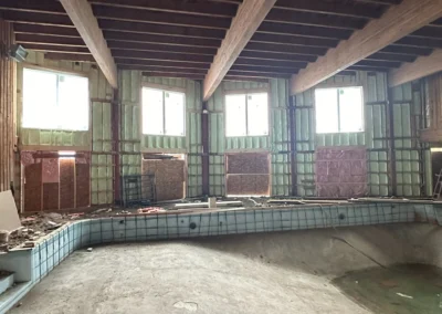 An empty indoor swimming pool is surrounded by unfinished walls with exposed framing and green spray foam insulation under a ceiling with large wooden beams