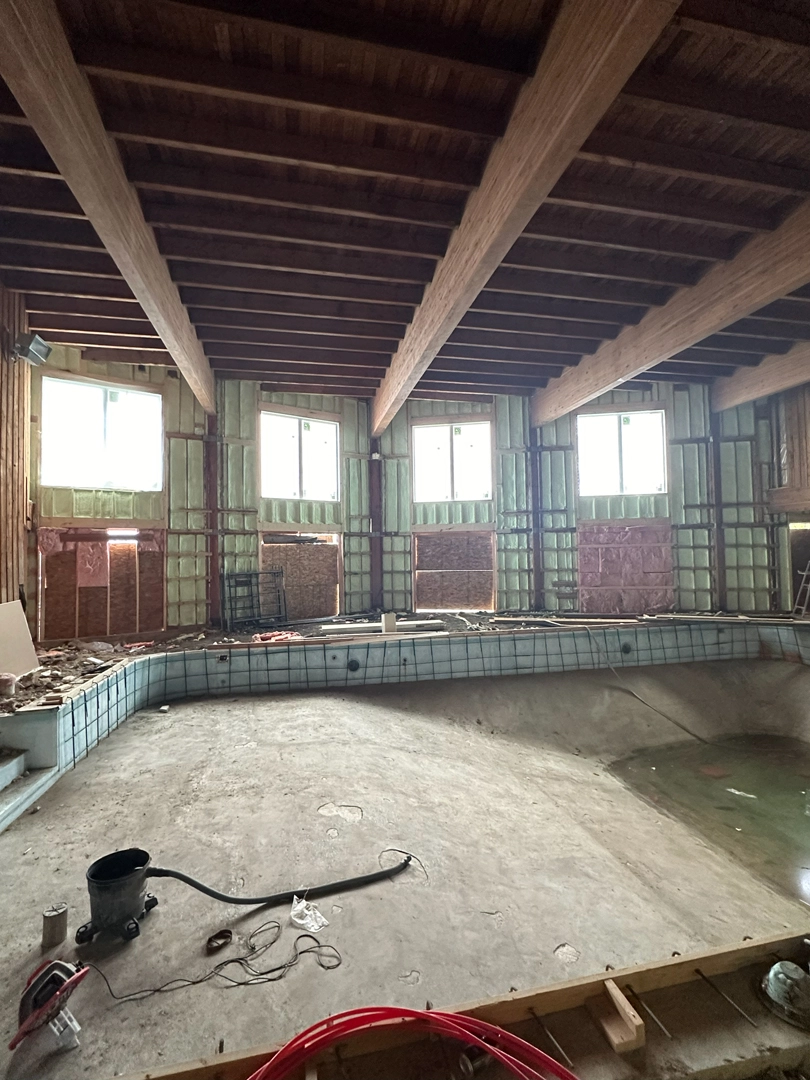 An empty indoor swimming pool is surrounded by unfinished walls with exposed framing and green spray foam insulation under a ceiling with large wooden beams