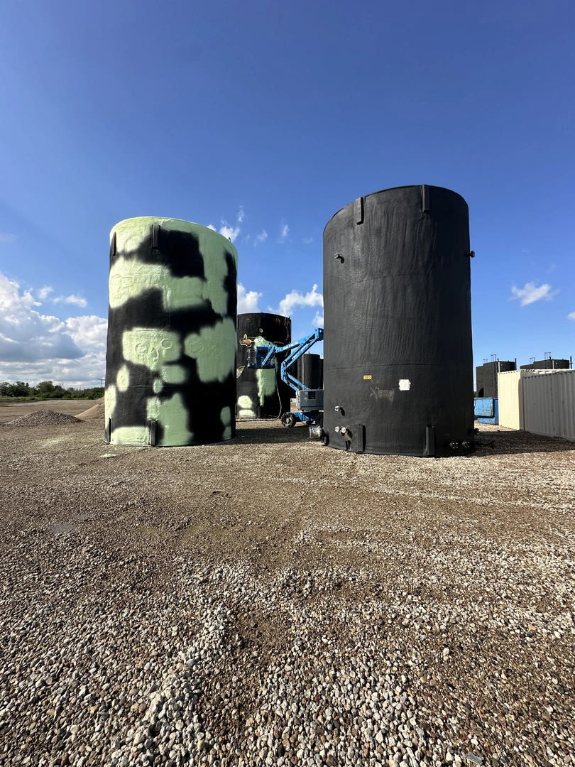 Three large cylindrical tanks, two black and one green with black splotches, sit on a gravel surface under a blue sky