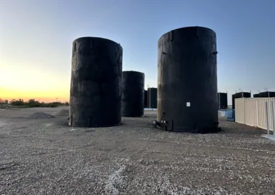 Three large, black cylindrical tanks sit on a gravel surface under a clear sky at sunset, with a faint horizon in the background