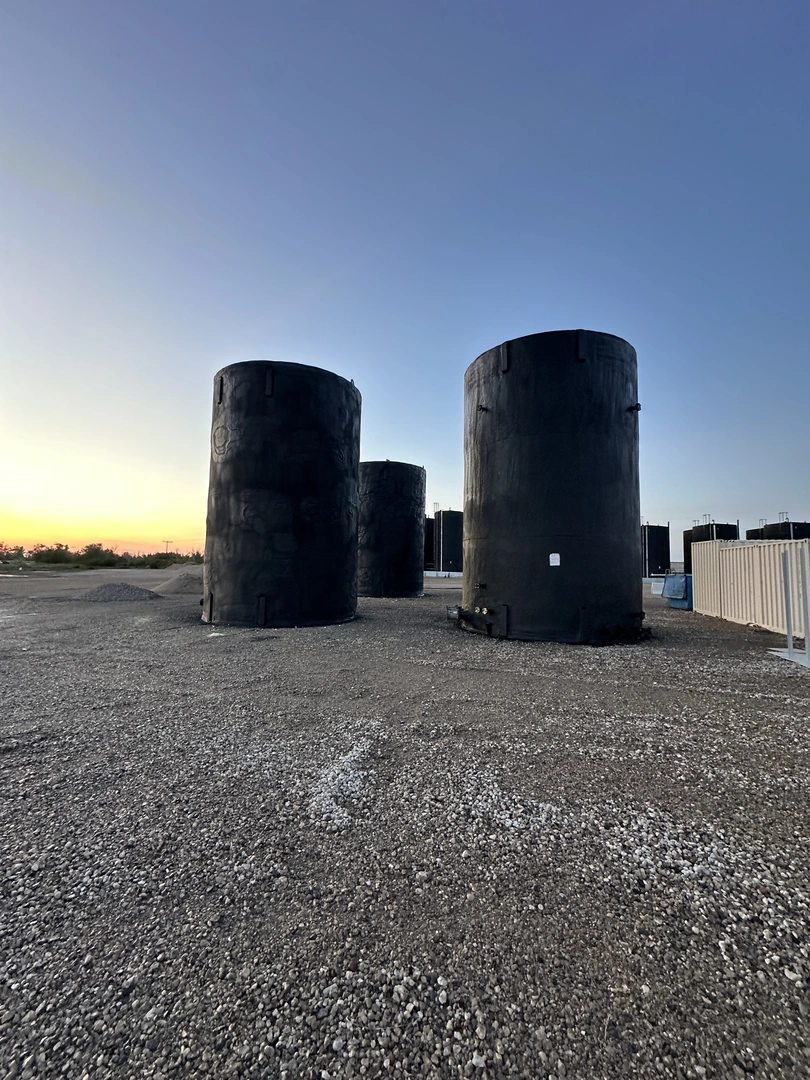Three large, black cylindrical tanks sit on a gravel surface under a clear sky at sunset, with a faint horizon in the background